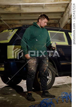 A man stands in a workshop, dressed in a green hoodie and paint-splattered pants. He holds a spray paint tool, preparing for touch-ups on a partially restored vehicle, with tools around him. 125417214
