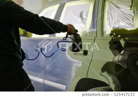 A worker focuses on applying paint to a vehicle using a spray gun in a dimly lit workshop. The environment showcases an atmosphere of craftsmanship and detail-oriented work. A worker focuses on applying paint to a vehicle using a spray gun in a dimly lit workshop. The environment showcases an atmosphere of craftsmanship and detail-oriented work. 125417215