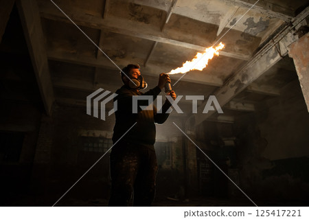 An individual in protective gear ignites a flamethrower inside a derelict structure, casting an orange glow in the darkened space. The environment evokes a sense of thrill and exploration. 125417221