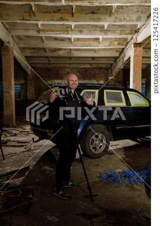 In a dimly lit, deserted industrial space, a photographer strikes a playful pose with his camera beside a parked black SUV, surrounded by rustic elements and scattered debris. In a dimly lit, deserted industrial space, a photographer strikes a playful pose with his camera beside a parked black SUV, surrounded by rustic elements and scattered debris. 125417226