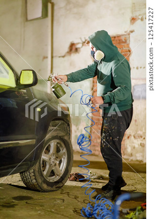 A person wearing a mask and a hoodie is painting a car in a garage. The individual uses a spray gun and is focused on achieving a smooth finish on the vehicle's surface. A person wearing a mask and a hoodie is painting a car in a garage. The individual uses a spray gun and is focused on achieving a smooth finish on the vehicle's surface. 125417227