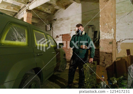 In a shadowy garage, a man wearing a gas mask applies green paint to an SUV, focused on transforming its appearance with a vibrant design in a hands-on project. 125417231