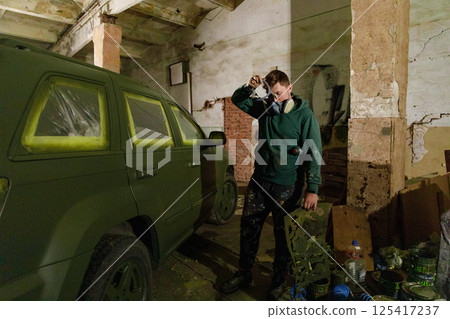 A person wearing a gas mask stands next to a partially painted green vehicle inside a warehouse. The ambient lighting and surroundings convey an atmosphere of urban artistry and transformation. 125417237
