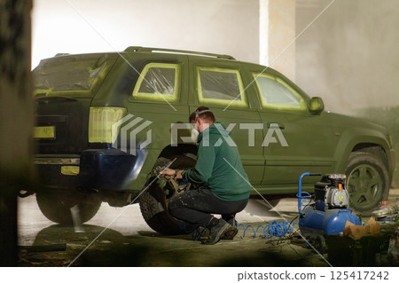 A man concentrates on painting a green SUV in a workshop. The surroundings are dimly lit with tools and paint visible, illustrating an engaging restoration process during the evening. 125417242