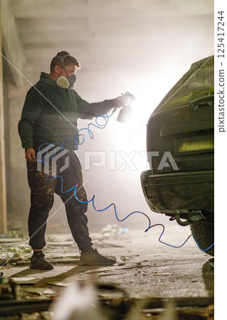 A person in a mask paints a car with spray paint in a dilapidated warehouse, surrounded by debris and a soft glow of light illuminating the scene, highlighting a moment of artistic expression. A person in a mask paints a car with spray paint in a dilapidated warehouse, surrounded by debris and a soft glow of light illuminating the scene, highlighting a moment of artistic expression. 125417244