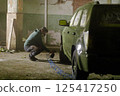 A worker is carefully painting a vehicle inside a dimly lit, abandoned warehouse. The setting features rough walls and scattered debris, highlighting the restoration effort underway. 125417250