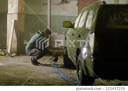 A worker is carefully painting a vehicle inside a dimly lit, abandoned warehouse. The setting features rough walls and scattered debris, highlighting the restoration effort underway. 125417250