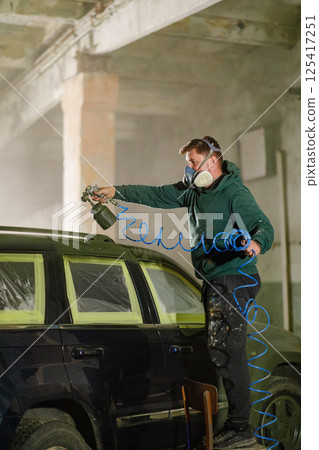 A man in a green hoodie wears a mask and goggles while using a spray gun to paint a vehicle inside a workshop. Protective tape covers parts of the car for precision work. 125417251