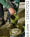 A person is holding a paint can, pouring vibrant green paint as they prepare for a creative endeavor in a dimly lit workshop. Various other paint cans are scattered around. 125417252