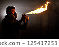 A man wearing a protective mask ignites an aerosol spray can, producing a powerful flame. The setting is dimly lit, enhancing the dramatic effect of the fire. 125417253