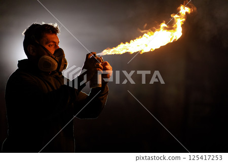 A man wearing a protective mask ignites an aerosol spray can, producing a powerful flame. The setting is dimly lit, enhancing the dramatic effect of the fire. 125417253