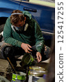 A young man, dressed in a green hoodie and protective gear, carefully pours paint onto the ground while crouched beside a parked vehicle. 125417255