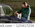 A man wearing a hoodie focuses intently on replacing windshield wipers on an SUV. The workshop has minimal lighting, creating a productive yet casual atmosphere. 125417256