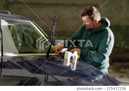 A man wearing a hoodie focuses intently on replacing windshield wipers on an SUV. The workshop has minimal lighting, creating a productive yet casual atmosphere. 125417256