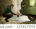 A craftsman carefully applies paint to the rear of a vehicle in a deserted warehouse, wearing a mask for protection. The workspace is filled with debris and shadows, highlighting his focused work. 125417259