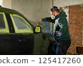A worker focuses on restoring a car in a workshop, applying paint with precision while wearing safety gear. The environment is filled with tools and projects underway, highlighting craftsmanship. 125417260