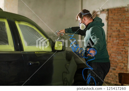 A worker focuses on restoring a car in a workshop, applying paint with precision while wearing safety gear. The environment is filled with tools and projects underway, highlighting craftsmanship. 125417260