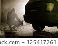 In a dimly lit urban area at night, an individual wearing a gas mask is crouching next to a vehicle, seemingly focused on a task involving tools and equipment. The atmosphere is tense and mysterious. 125417261