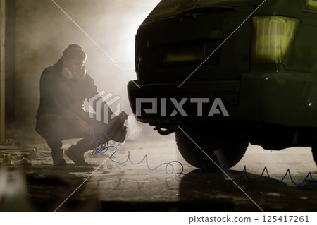 In a dimly lit urban area at night, an individual wearing a gas mask is crouching next to a vehicle, seemingly focused on a task involving tools and equipment. The atmosphere is tense and mysterious. 125417261