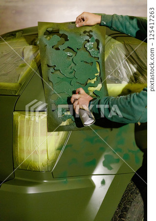 A person applies a green camouflage design on a vehicle using a spray can and a stencil. The workshop setting shows preparations for painting, highlighting creativity and craftsmanship. A person applies a green camouflage design on a vehicle using a spray can and a stencil. The workshop setting shows preparations for painting, highlighting creativity and craftsmanship. 125417263