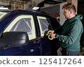 A man in a green hoodie meticulously applies masking tape around the windows of a dark blue SUV in a workshop. The process occurs in an industrial setting under dim lighting. 125417264