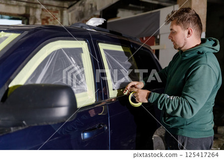 A man in a green hoodie meticulously applies masking tape around the windows of a dark blue SUV in a workshop. The process occurs in an industrial setting under dim lighting. A man in a green hoodie meticulously applies masking tape around the windows of a dark blue SUV in a workshop. The process occurs in an industrial setting under dim lighting. 125417264
