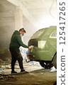 A man wears a mask while spray painting a vehicle inside a dimly lit workshop filled with dust and debris. He focuses on the car's rear, engaged in a detailed restoration project. 125417265