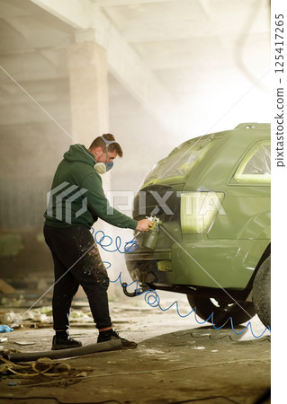 A man wears a mask while spray painting a vehicle inside a dimly lit workshop filled with dust and debris. He focuses on the car's rear, engaged in a detailed restoration project. 125417265
