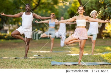 Multinational yoga enthusiasts with female coach perform vrikshasana in park Multinational yoga enthusiasts with female coach perform vrikshasana in park 125418853