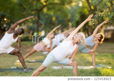 Elderly woman practices yoga pose Anjaneyasana with other people of different nationalities in summer park Elderly woman practices yoga pose Anjaneyasana with other people of different nationalities in summer park 125418925