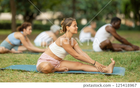 Girl stretching in Janu Sirsasana at group yoga class in park Girl stretching in Janu Sirsasana at group yoga class in park 125418967