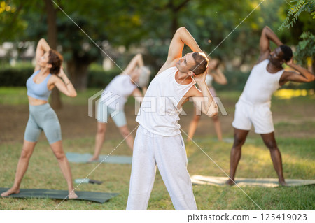 Man performing Bending Tree Pose during group yoga in park 125419023