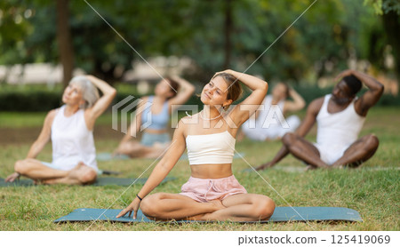 Multinational yoga enthusiasts with female coach perform padmasana in park Multinational yoga enthusiasts with female coach perform padmasana in park 125419069