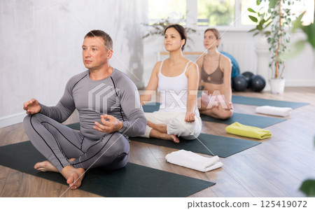 Positive man in sports clothes having yoga class in group sitting in lotus pose in yoga studio Positive man in sports clothes having yoga class in group sitting in lotus pose in yoga studio 125419072