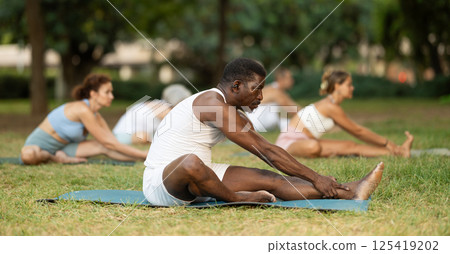 Multinational yoga enthusiasts with African male coach perform Janu Shirshasana in park Multinational yoga enthusiasts with African male coach perform Janu Shirshasana in park 125419202