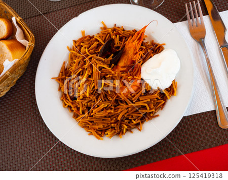 Fideua marinera(Spanish) seafood noodles with prawns and mussels served with sauce on a white plate in a restaurant Fideua marinera(Spanish) seafood noodles with prawns and mussels served with sauce on a white plate in a restaurant 125419318