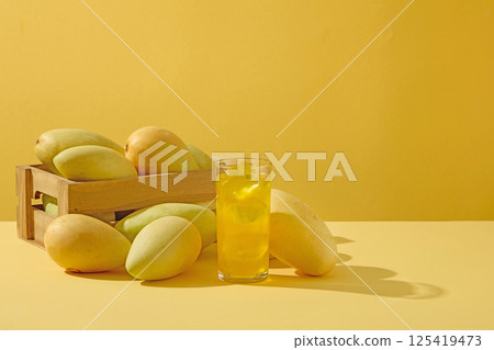 Freshly harvested mangoes placed in a rectangular wooden crate. Mango juice with ice served in a glass, surrounded by ripe mangoes of different sizes. Shadows of the mangoes cast on the surface. 125419473