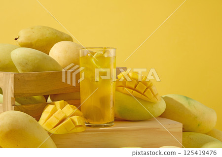 Glass of mango juice and clear ice cubes. Half of a mango with slices dividing the mango flesh into cubes, curved and placed on others. The mangifera indica are placed around the wooden crate. Glass of mango juice and clear ice cubes. Half of a mango with slices dividing the mango flesh into cubes, curved and placed on others. The mangifera indica are placed around the wooden crate. 125419476
