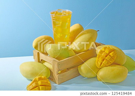 The plastic cup of mango juice has a slight condensation mark. Ripe mangoes are displayed around a rustic wooden tray, with a bright yellow color and smooth skin. Presented on a blue background. The plastic cup of mango juice has a slight condensation mark. Ripe mangoes are displayed around a rustic wooden tray, with a bright yellow color and smooth skin. Presented on a blue background. 125419484