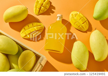 Whole mangoes, halved and diced, scattered around on an orange background. The glass bottle contains pure juice from fresh Mahachanok Mangoes, the juice has a beautiful bright yellow color. Whole mangoes, halved and diced, scattered around on an orange background. The glass bottle contains pure juice from fresh Mahachanok Mangoes, the juice has a beautiful bright yellow color. 125419491