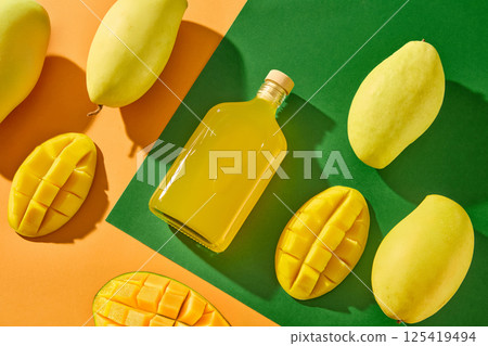 Composition of mangoes and halved mango slices placed in straight rows, parallel to a glass bottle containing pure juice. Unlabeled glass bottle for advertising. Unique green and orange background. 125419494
