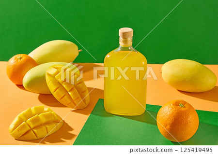 A glass bottle of juice is placed in the center, surrounded by ripe mangoes. The mangoes are cut in half and the flesh is scored into squares. A few oranges with rough skin are scattered around. 125419495