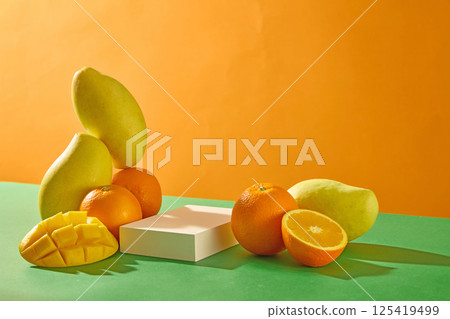 Mangoes are stacked vertically on top of round oranges, surrounded by mango slices with evenly spaced squares. A rectangular white pedestal is placed between the fruits. Empty space for text.  125419499
