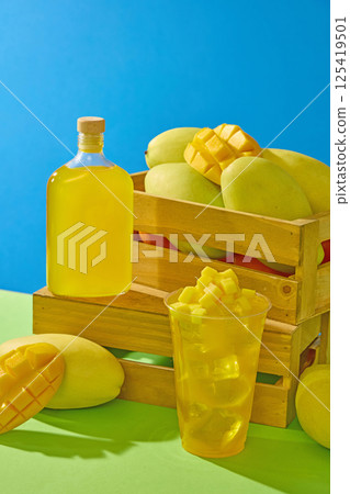 Vertical photo of wooden crates filled with several ripe mangifera indica. Plastic cups of mango juice with diced mangoes decorated on top. Glass pitcher placed on the edge of a wooden crate. 125419501