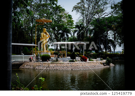 Singburi, Thailand, March 23, 2025, Golden statue of serene figure beside a reflective pond in Wat Phikun Thong, amidst lush greenery 125419779