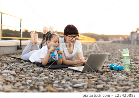 Mother and daughter enjoying outdoor activity watching laptop fitness course using dumbbells and yoga mat Mother and daughter enjoying outdoor activity watching laptop fitness course using dumbbells and yoga mat 125420882