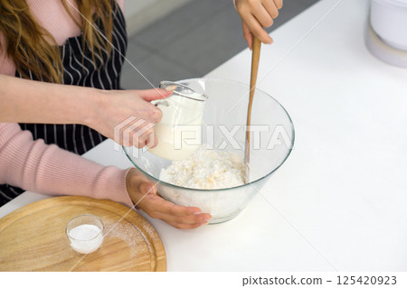 Two women make dessert in the kitchen. One people stirring a white cake batter or icing mixture. The other pouring fresh milk in a clear glass bowl. Two women make dessert in the kitchen. One people stirring a white cake batter or icing mixture. The other pouring fresh milk in a clear glass bowl. 125420923