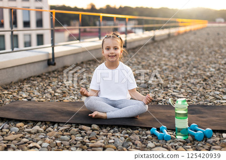 Child practicing meditation sitting cross-legged on mat at rooftop sunrise with water and weights. Causal fitness concept emphasizing health, wellness, early age motivation. Child practicing meditation sitting cross-legged on mat at rooftop sunrise with water and weights. Causal fitness concept emphasizing health, wellness, early age motivation. 125420939