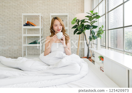 Young asian woman enjoy morning coffee or tea in mug on comfortable bed in a bright, cozy bedroom at home. Lifestyle Concept 125420973