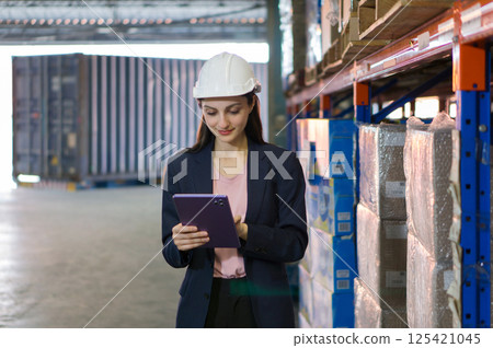 Young business woman in suit and safety helmet review inventory record on tablet computer. Stand among stacked boxes in a busy storage area. Working in container stuffing and cargo securing service. 125421045